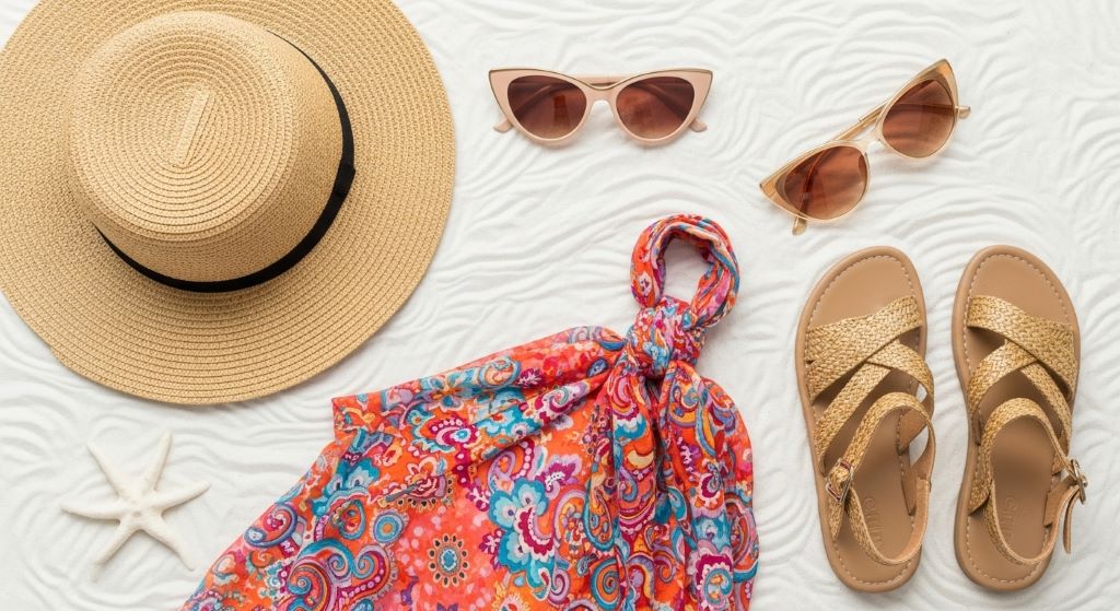 Flat lay of women’s beach summer outfit with a straw hat, cat-eye sunglasses, sarong, and sandals on white sand