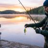 An angler holding a colorful fishing lure over a calm lake at sunrise, preparing to cast