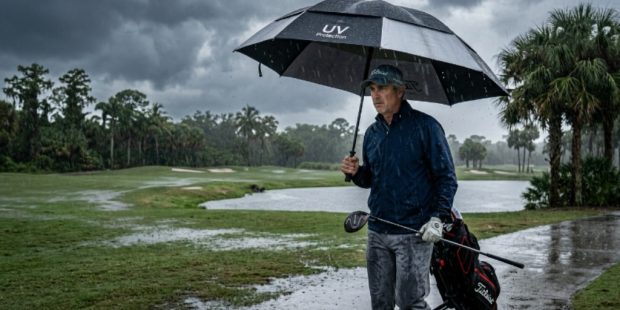 A golfer holding a large UV golf umbrella on a Florida course during a heavy afternoon rainstorm