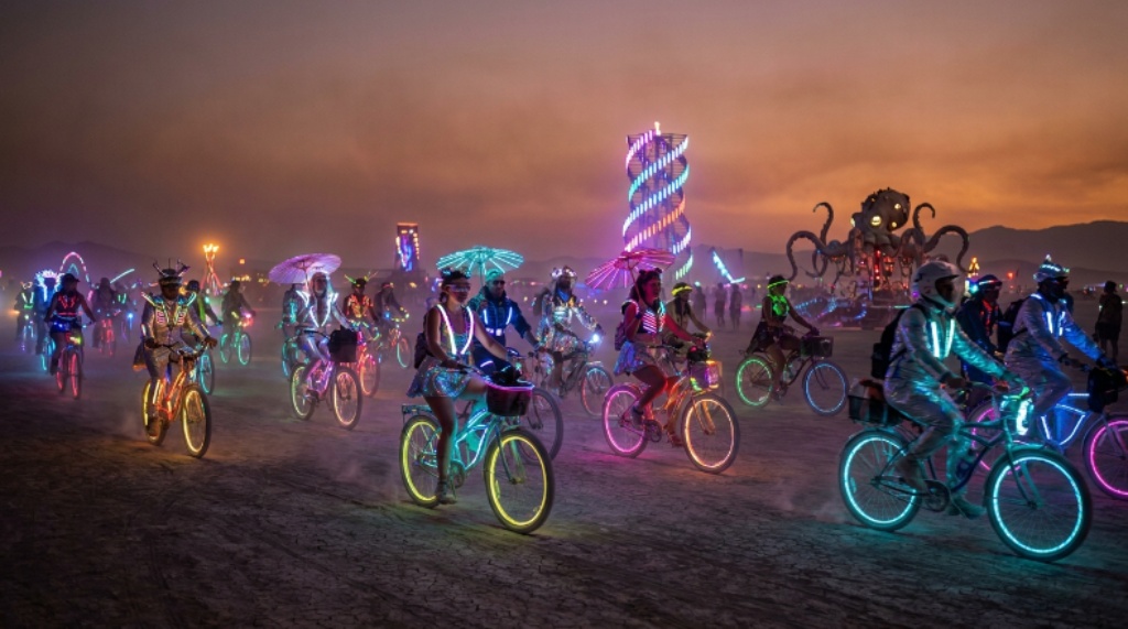 Participants in elaborate cosmic costumes riding decorated, LED-lit bikes across the dusty playa at Burning Man 2026, with large-scale art installations glowing in the background