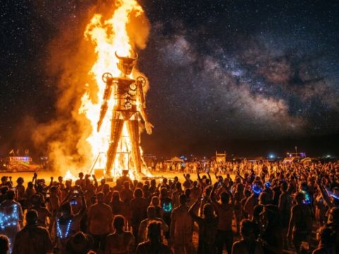 A massive wooden effigy surrounded by towering flames at night during Burning Man 2026 in Nevada's Black Rock Desert, with thousands of participants watching under a star-filled sky