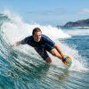 Surfer riding a blue ocean wave using a wooden bodysurfing handplane.